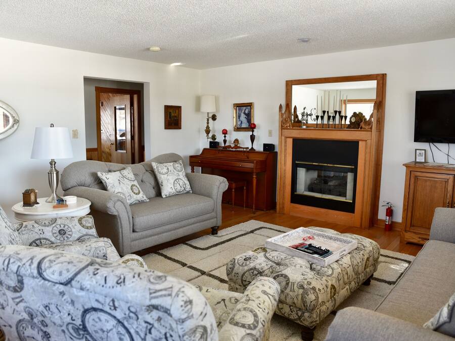 Uma sala de estar com sofá, pufê, poltrona, lareira, televisão e piano.
