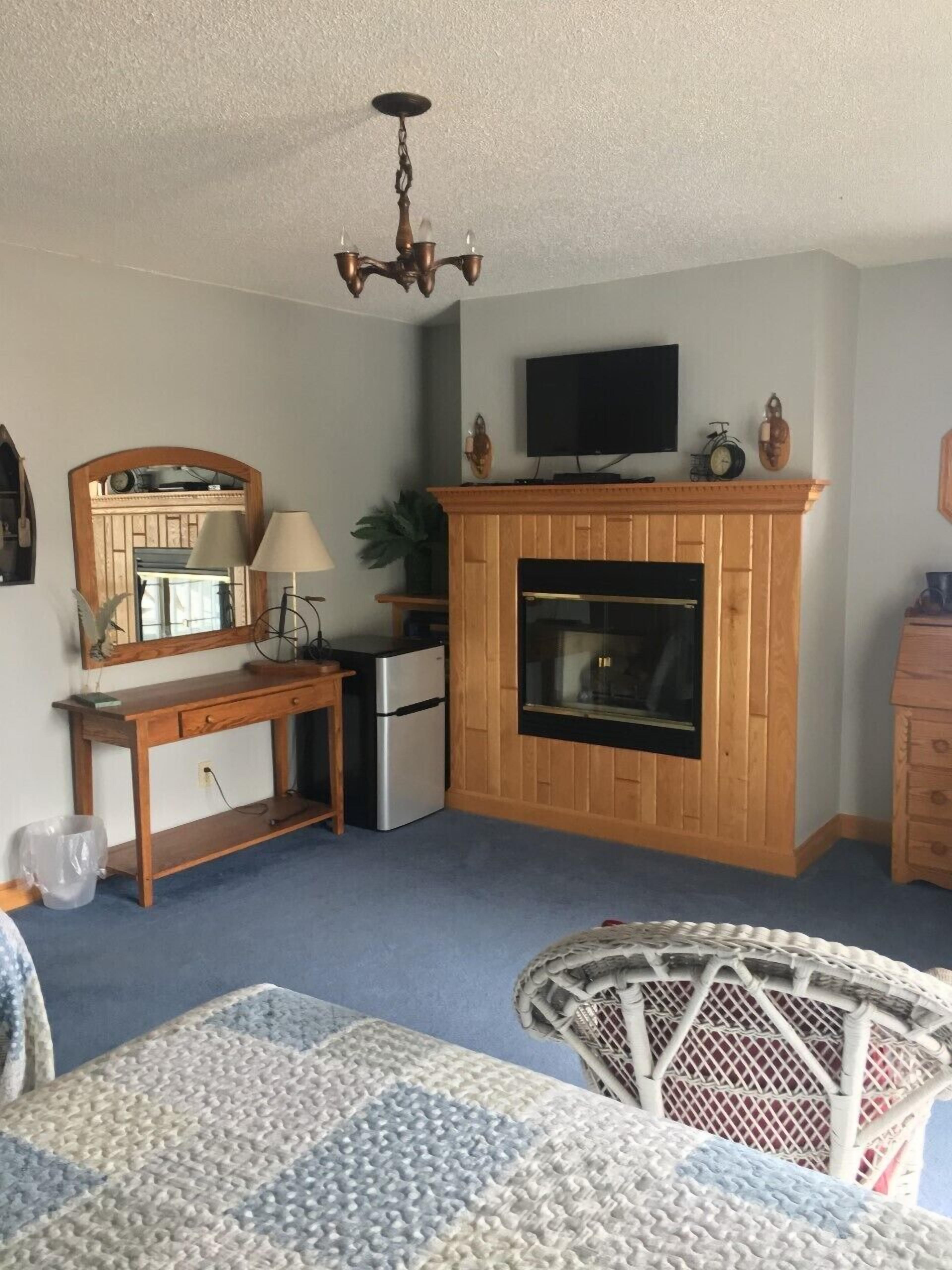 A cozy living room with a wooden fireplace, a television mounted above it, a small desk with a lamp, and a wicker chair.