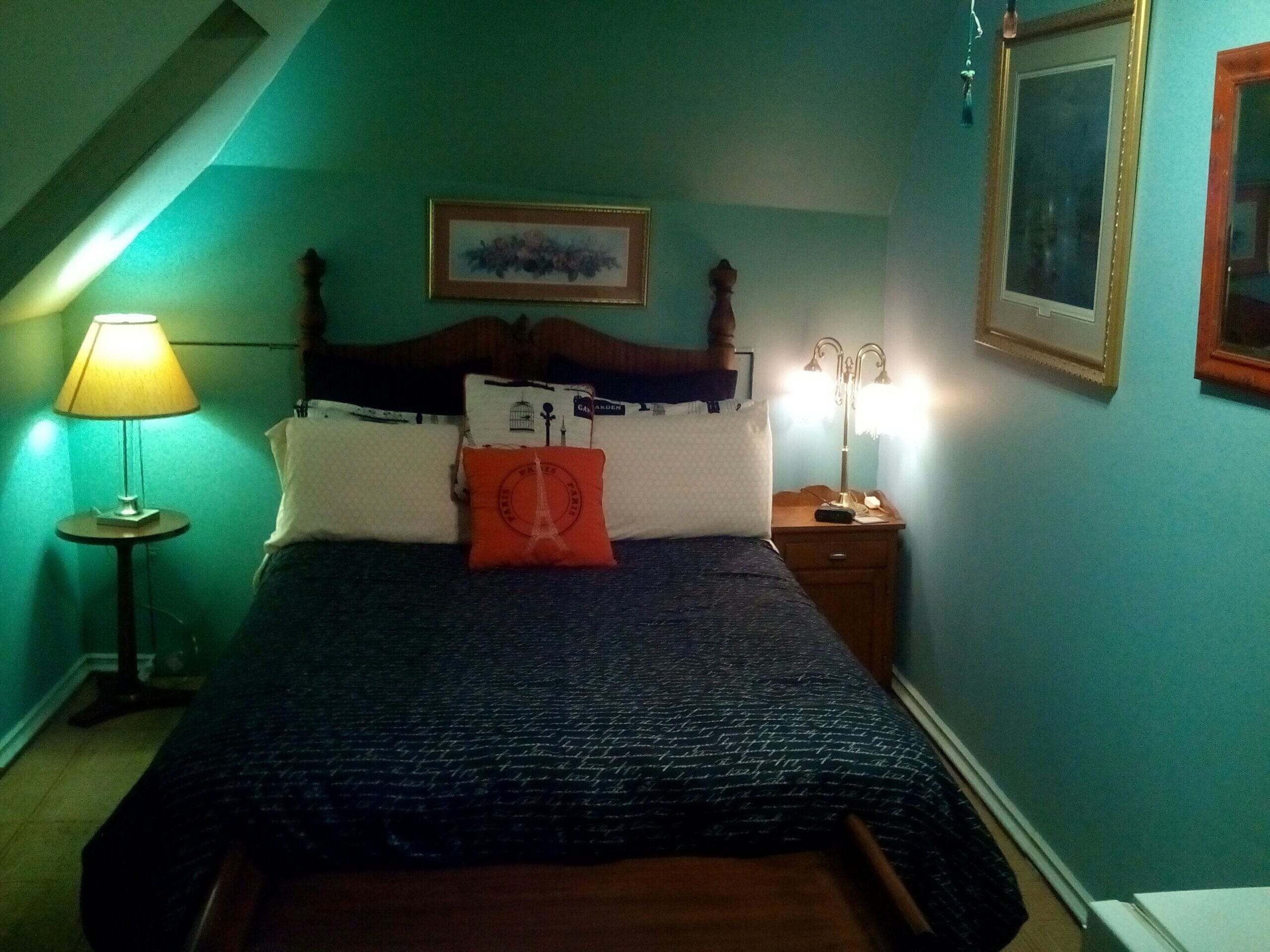 A neatly made bed with a dark patterned bedspread, a wooden headboard, and a single orange pillow. There are two bedside lamps on either side of the bed, and a framed picture hangs above the headboard.