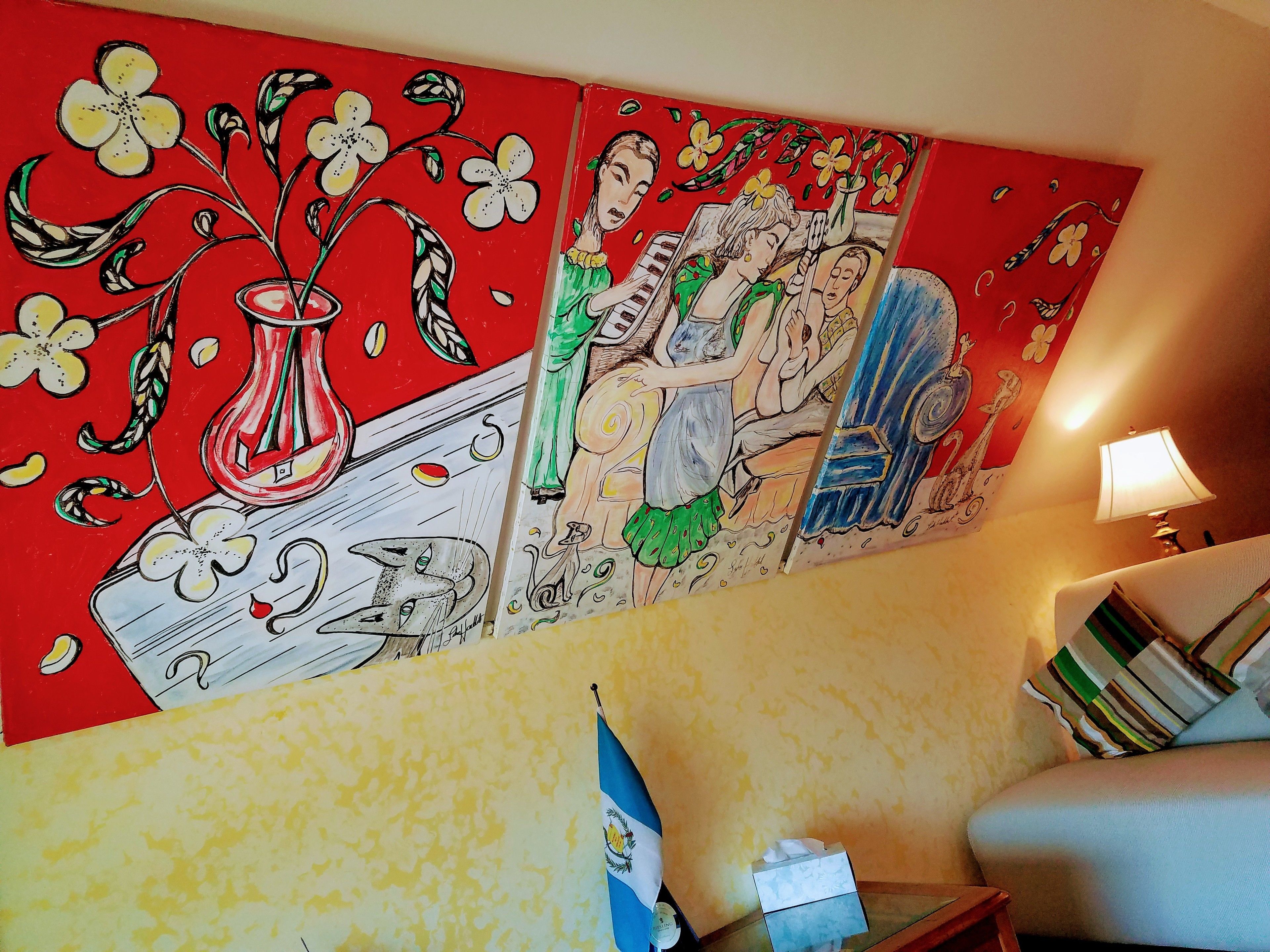 Una habitación con un cuadro grande y colorido en la pared, una bandera y una lámpara sobre una mesita auxiliar.