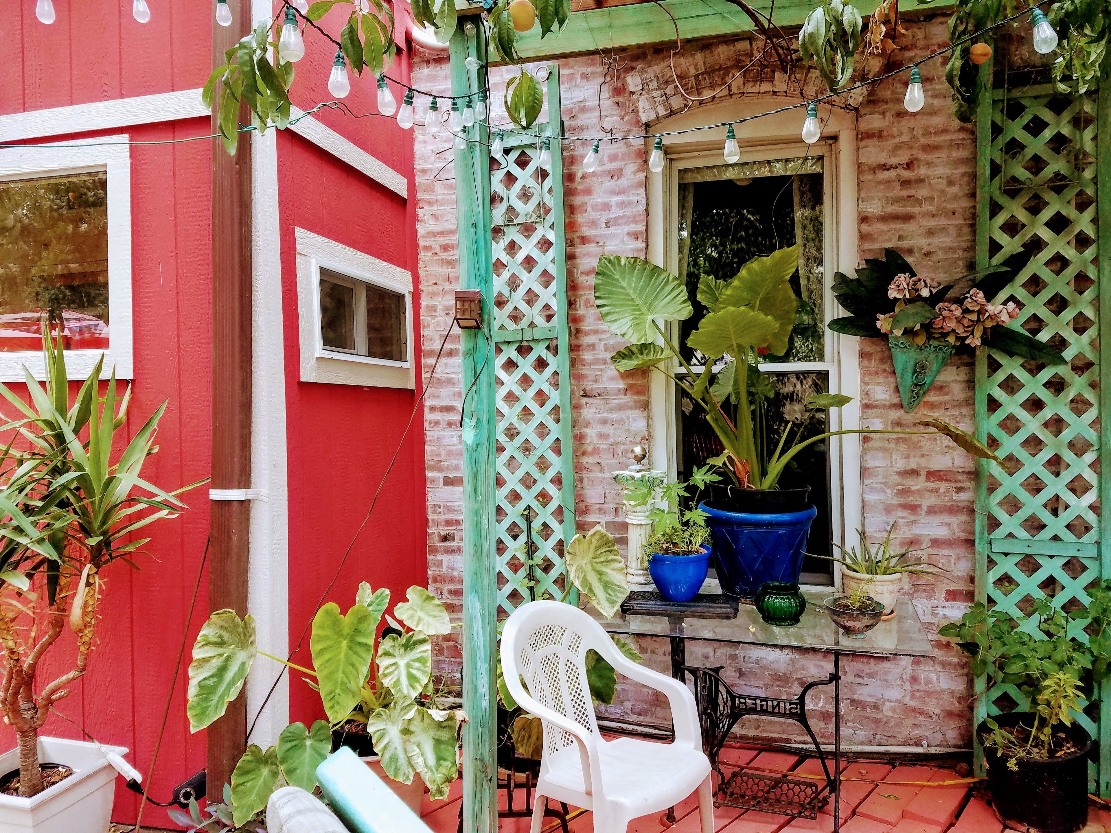 Um edifício vermelho com parede de tijolos, uma janela e uma porta. Há plantas em vasos e uma cadeira de plástico branca na parte externa.