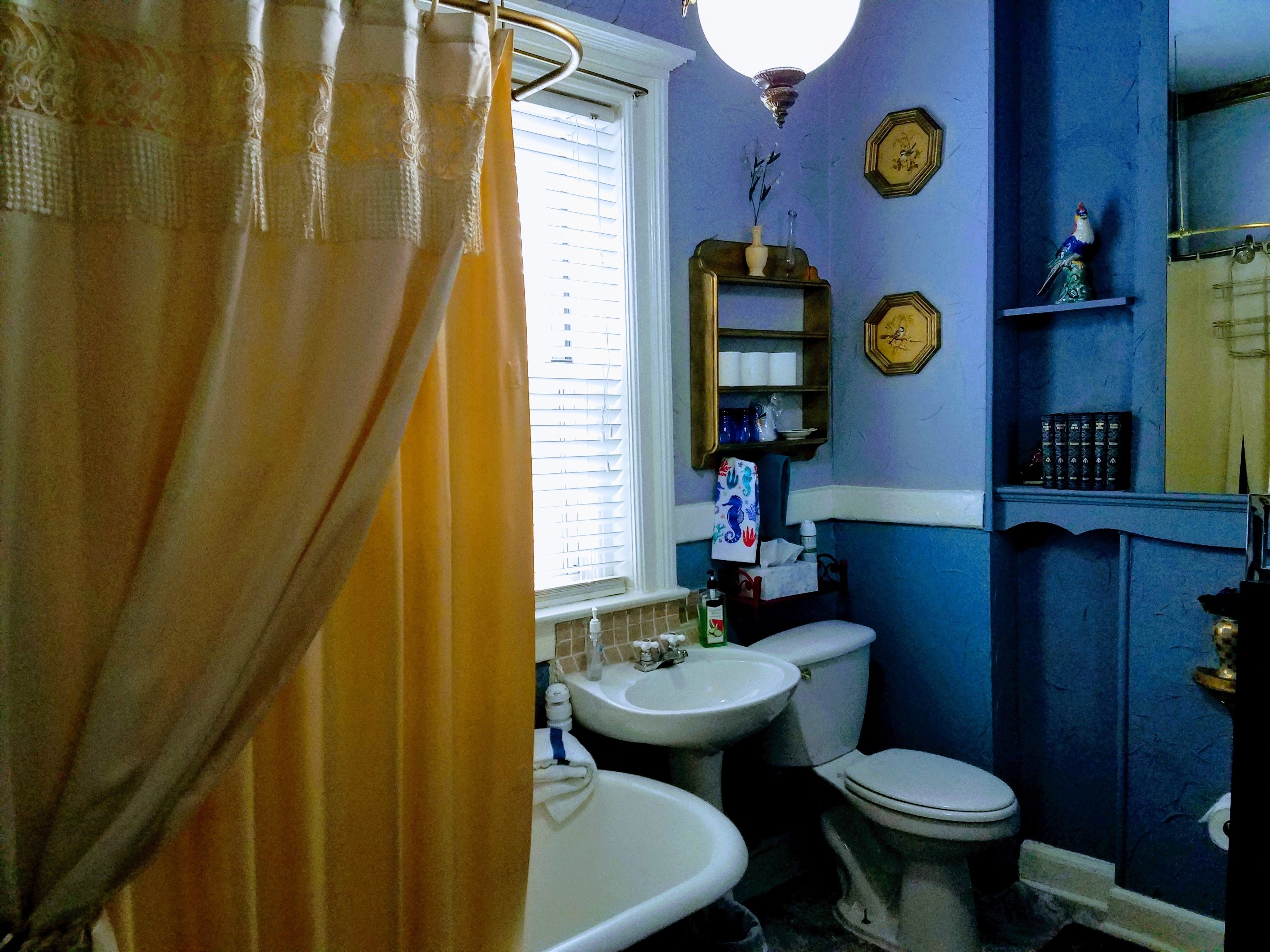 Uma casa de banho com banheira, sanita e lavatório. Há uma janela com persianas, um porta-toalhas e uma prateleira com artigos de higiene pessoal.