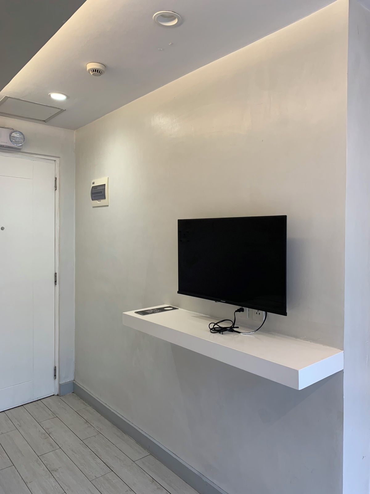 Un pasillo con una puerta blanca, un televisor montado en un estante blanco e iluminación empotrada en el techo.