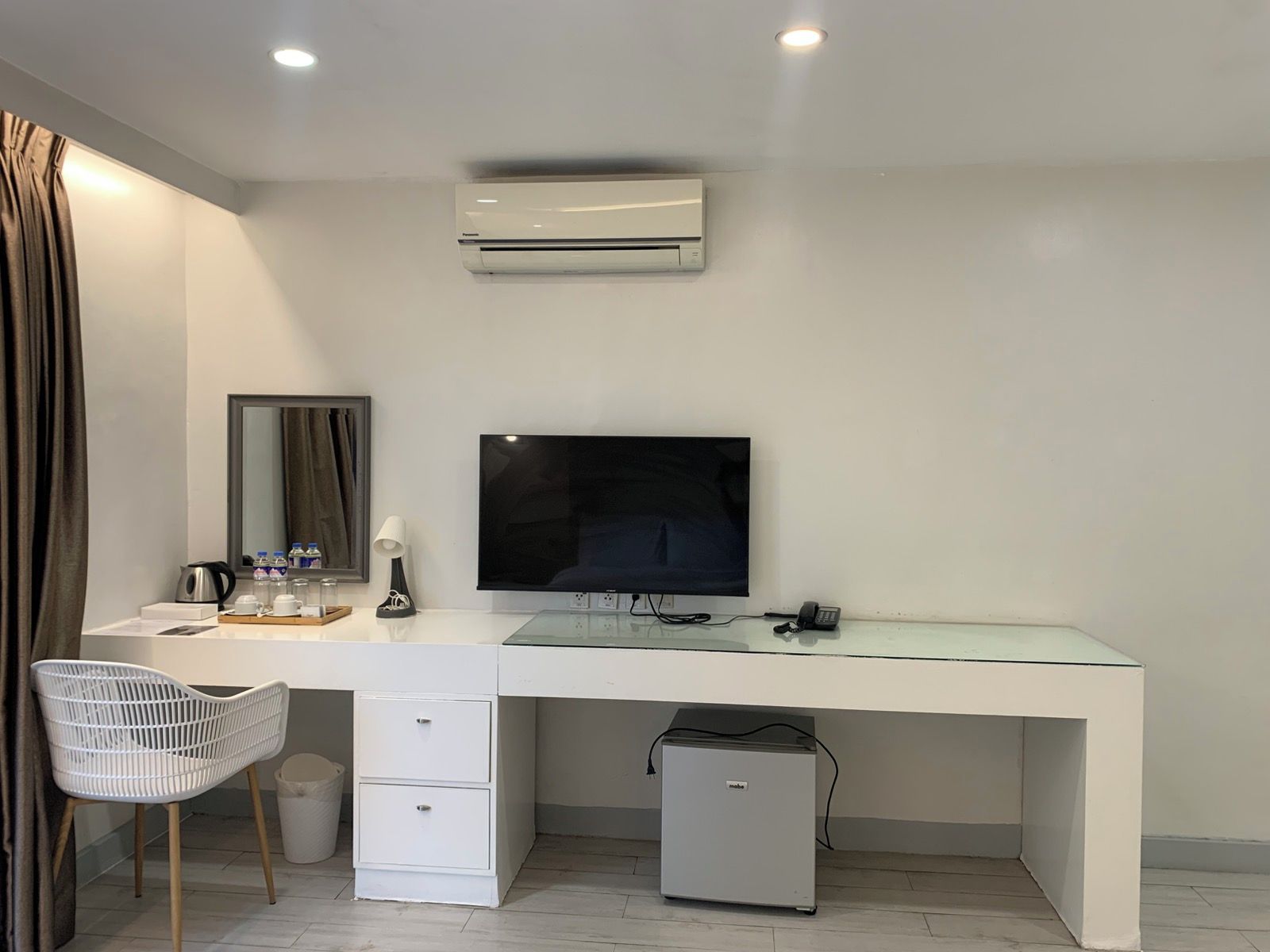 A modern hotel room with a white desk, a flat-screen TV, a small refrigerator, and a white chair.