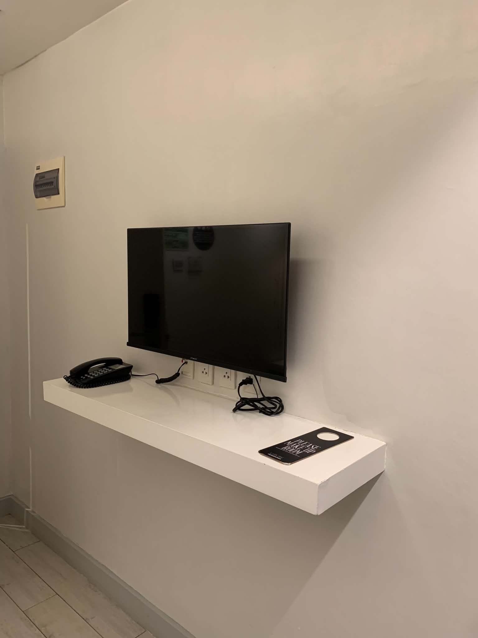 A flat-screen TV mounted on a white wall, with a telephone and a sign on a shelf below.