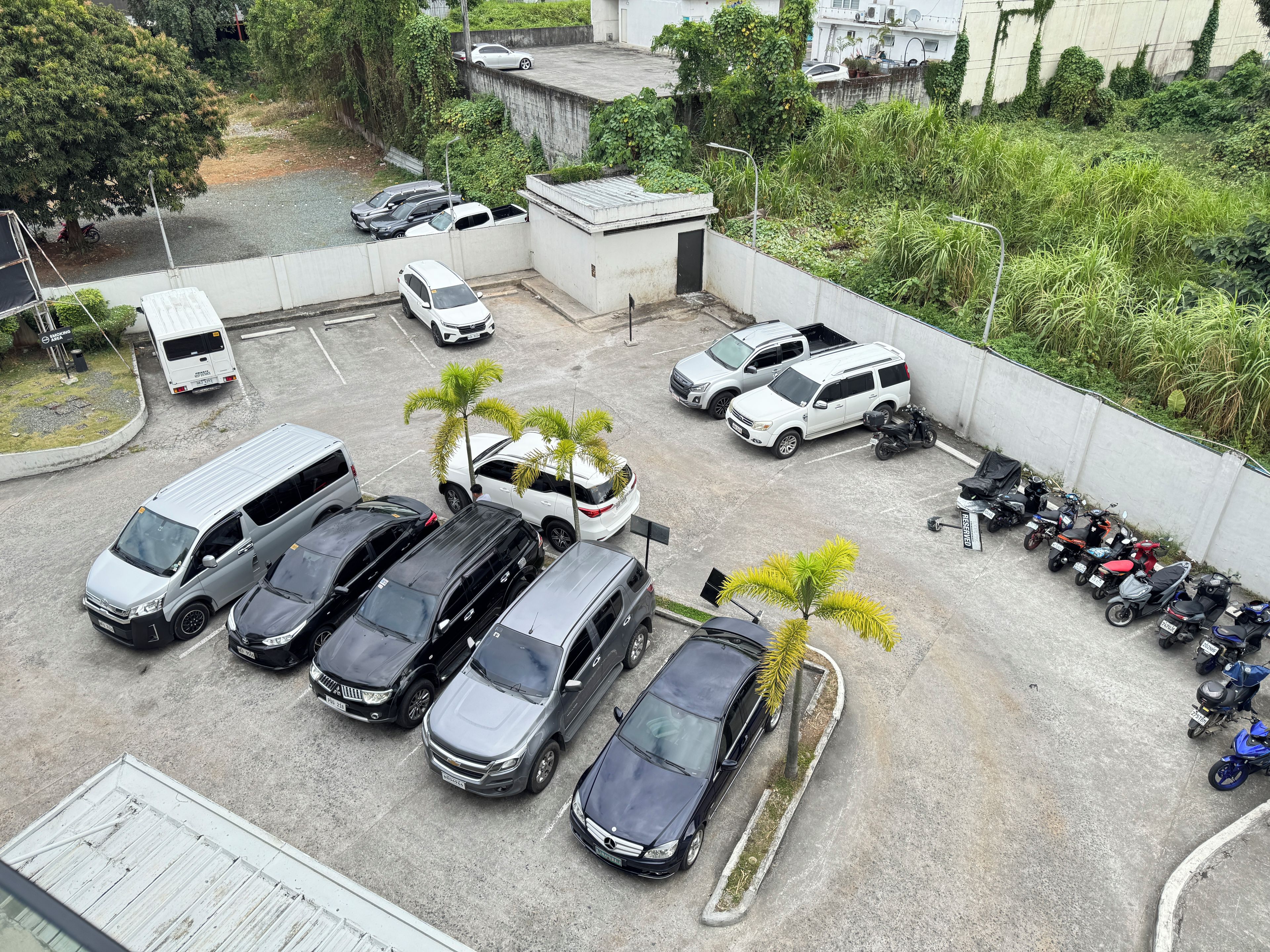 A parking lot with multiple cars and motorcycles, surrounded by greenery and a concrete wall.