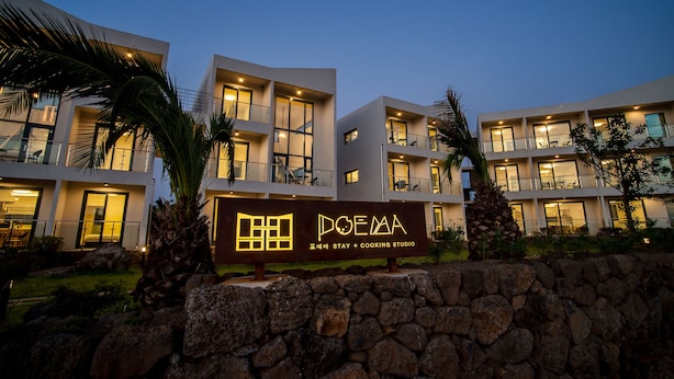 A modern hotel with a sign reading "DOEMA Stay + Cooking Studio." It features multiple balconies and a stone wall in the foreground. - Poema Stay (Jeju City)