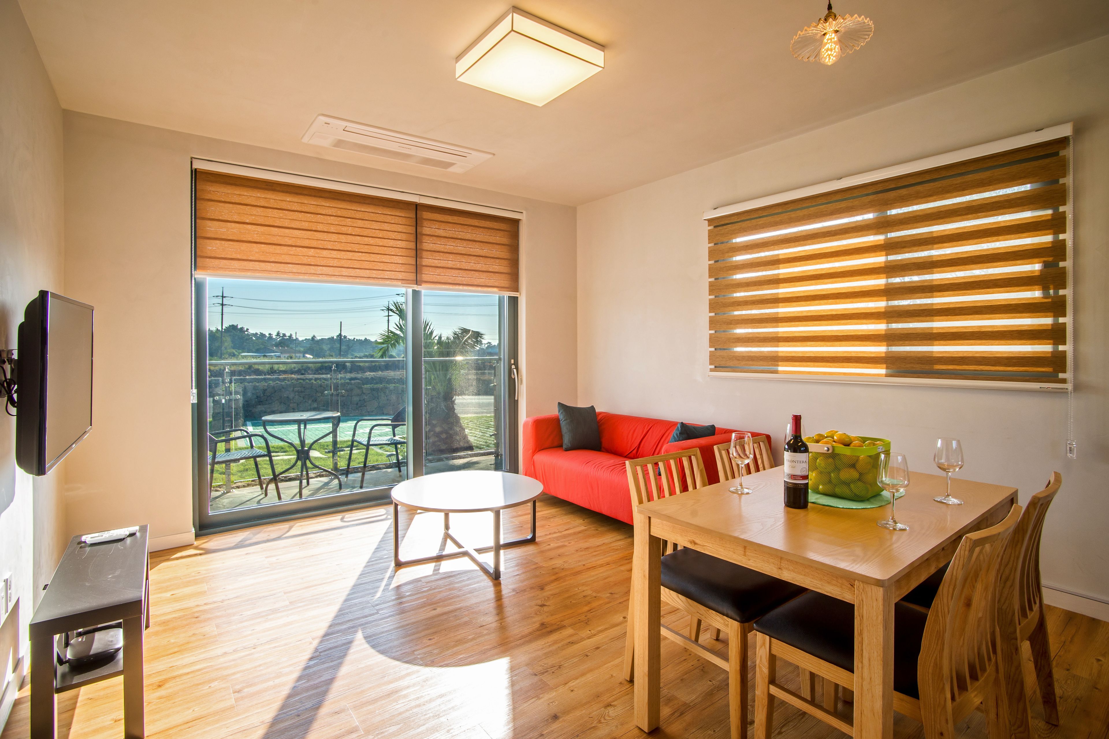 Un salon moderne avec un plancher en bois, un canapé rouge, une table à manger avec des chaises, un téléviseur et une grande fenêtre avec des stores.