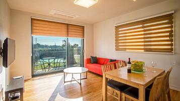 Un salon moderne avec un plancher en bois, un canapé rouge, une table à manger avec des chaises, un téléviseur et une grande fenêtre avec des stores.