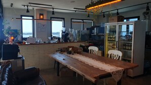 Un restaurante acogedor con pared de ladrillo, mesa y sillas de madera. Hay un refrigerador con botellas, una máquina de café y vistas al mar a través de amplias ventanas.
