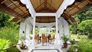 A covered outdoor seating area with a thatched roof, surrounded by lush greenery and a small pond. - Bali Hidden Paradise Villa (Seminyak)