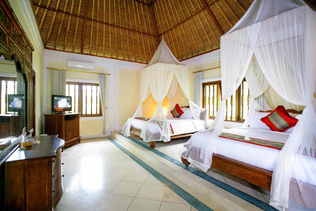 A hotel room with a thatched roof, two beds with white canopies, a wooden desk, and a television.