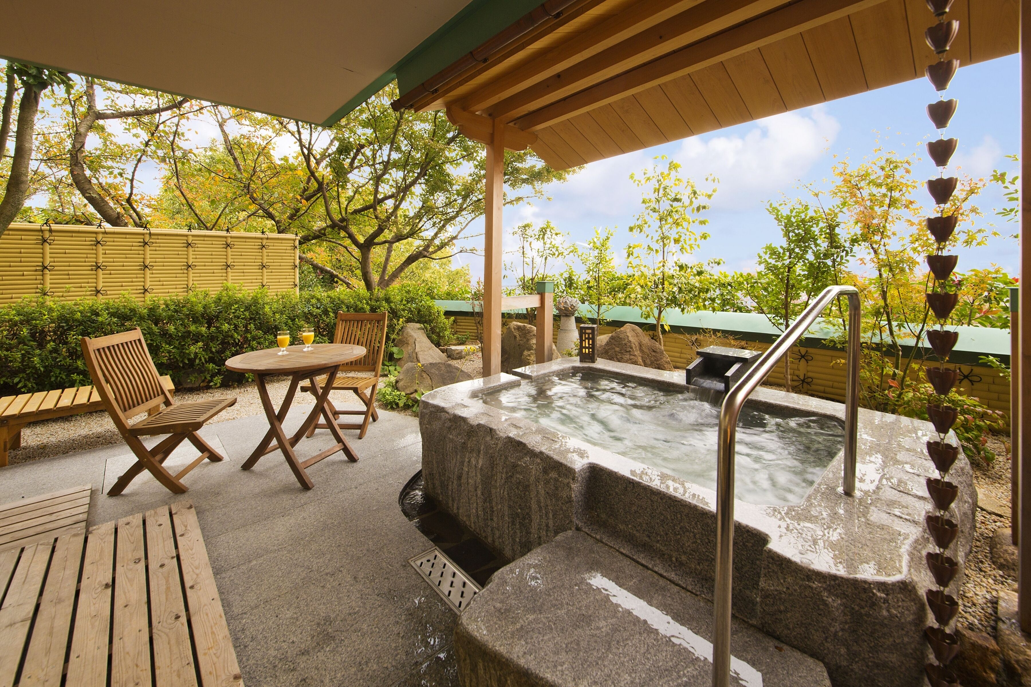 A hot tub area with a wooden table and chairs, surrounded by a stone wall and greenery.