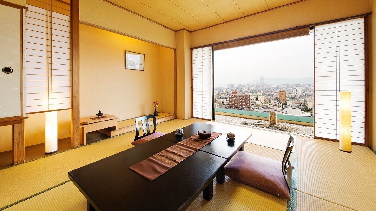 A traditional Japanese room with tatami flooring, a low table, and a view of a cityscape through large windows.