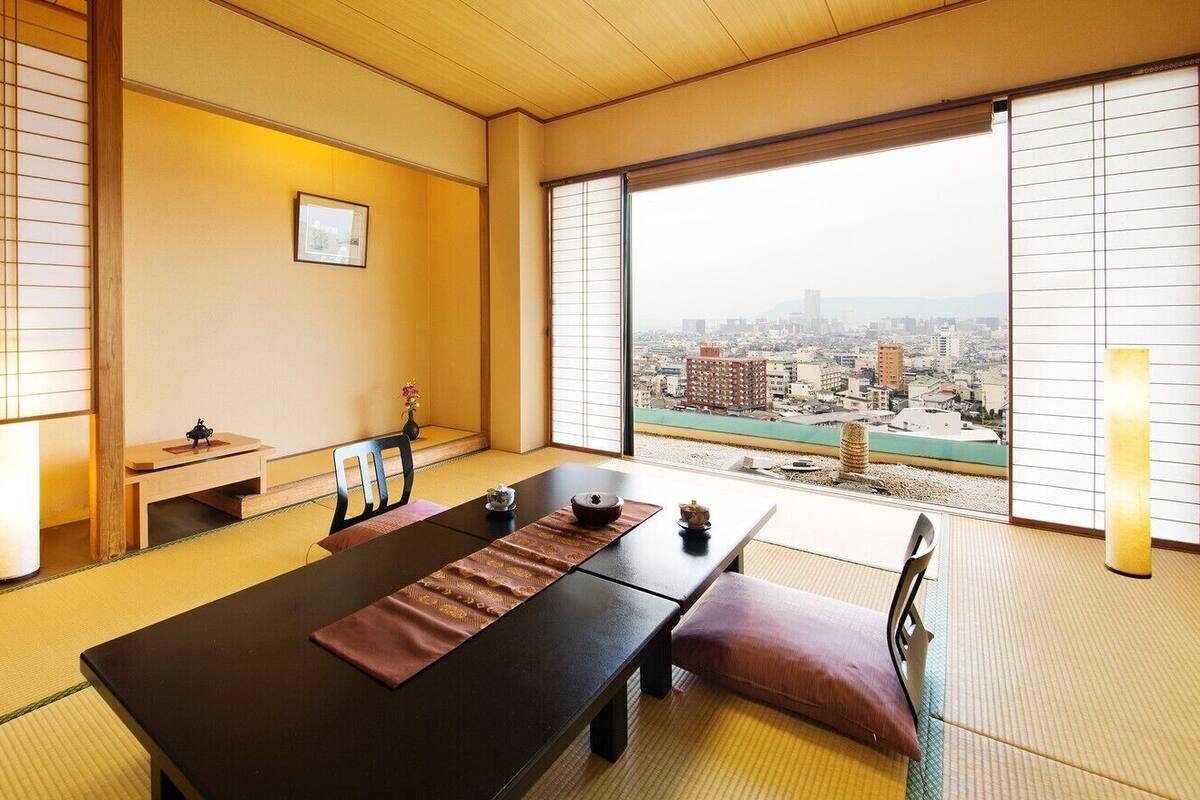 A traditional Japanese room with tatami flooring, a low table, and a view of a cityscape through large windows.