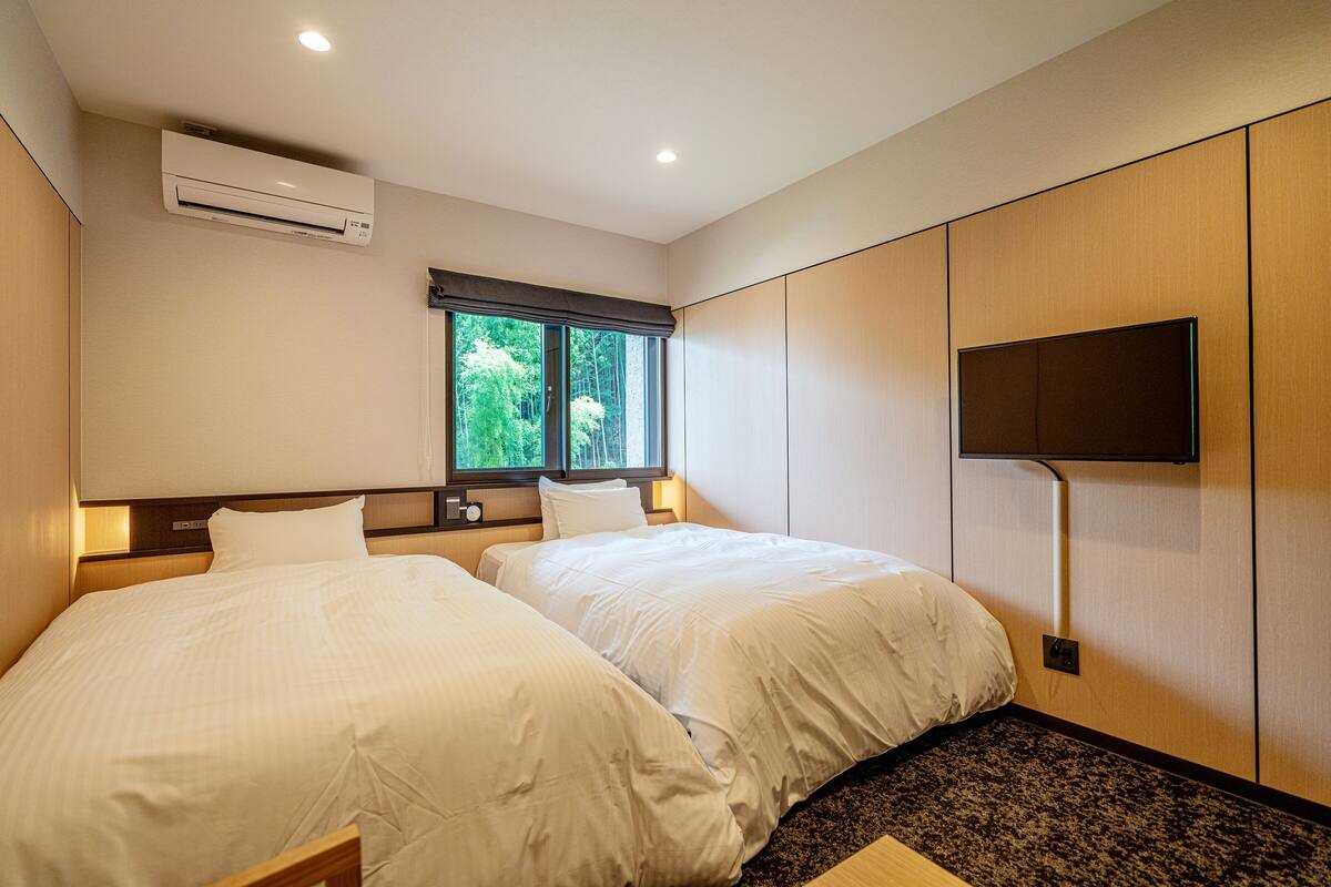 Habitación de hotel con dos camas, un equipo de aire acondicionado, una ventana con vistas a zonas verdes y un televisor de pantalla plana montado en la pared.
