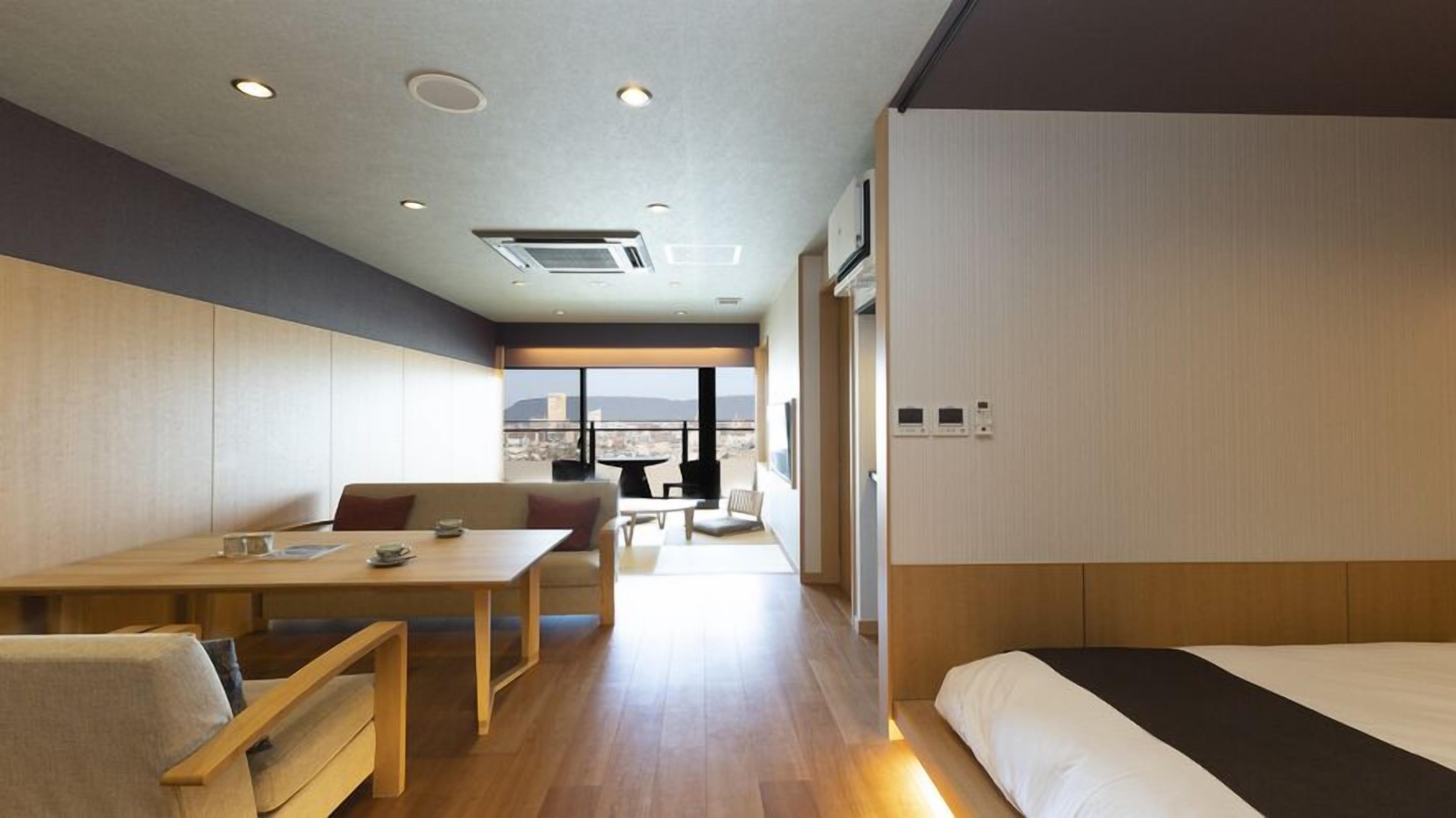 A modern hotel room with a wooden floor, a bed, a desk, and a balcony with a city view.