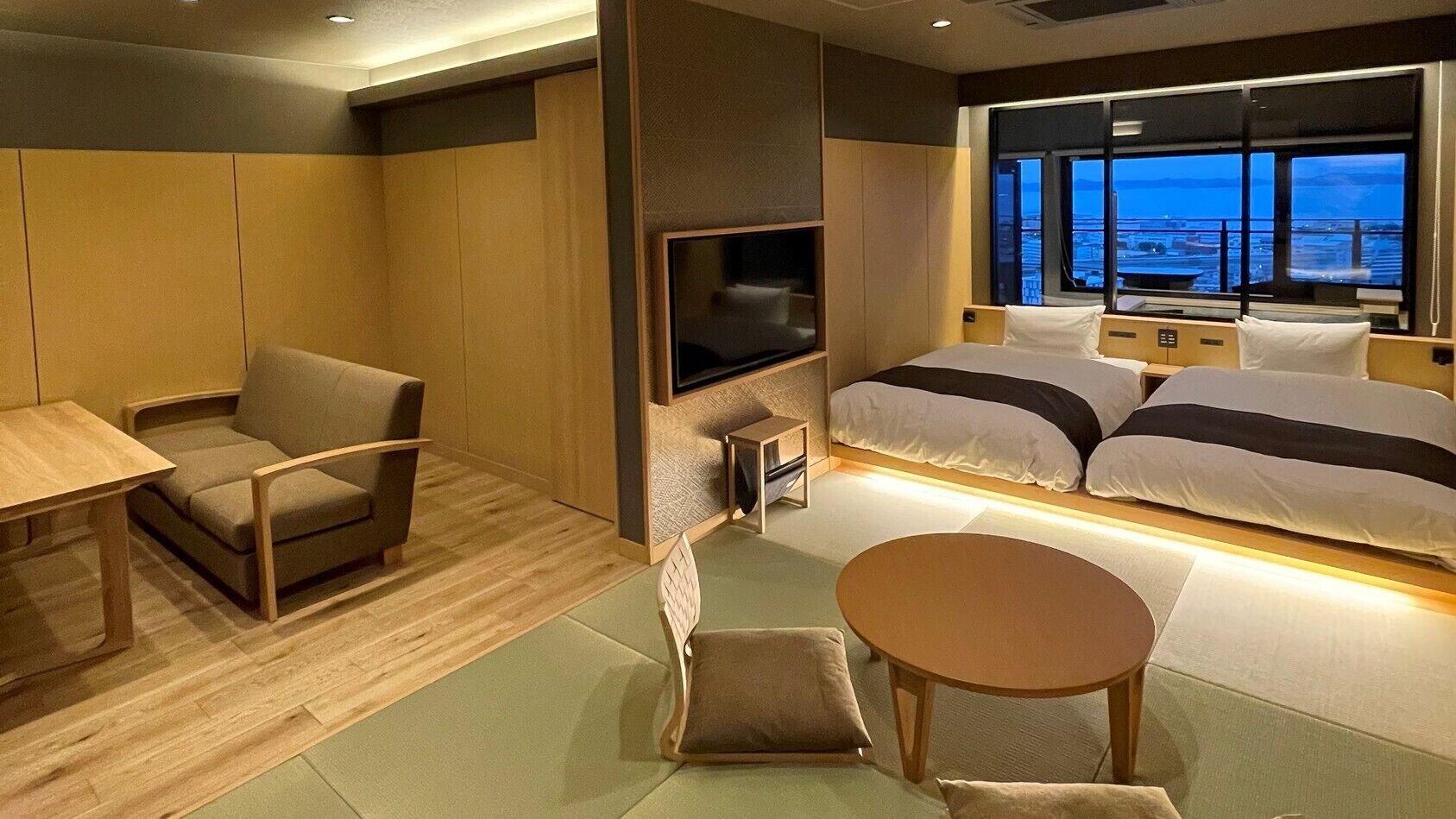 A hotel room with a bed, a TV, a seating area with a table, and a view of the city.