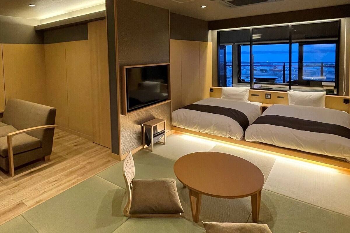 Habitación de hotel con cama, televisor, zona de estar con mesa y vista a la ciudad.