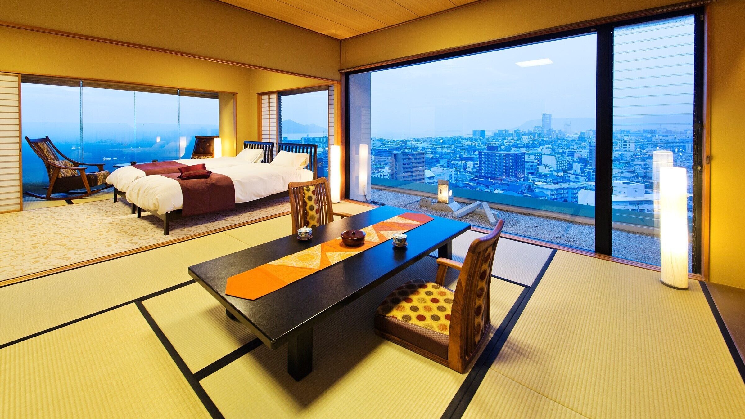 A hotel room with tatami flooring, a dining table, a sofa, and a large window with a city view.