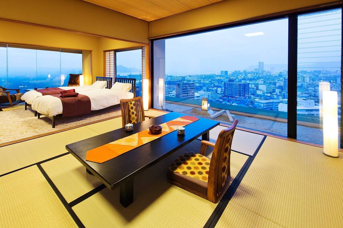 Habitación de hotel con suelo de tatami, una mesa de comedor, un sofá y un ventanal con vistas a la ciudad.