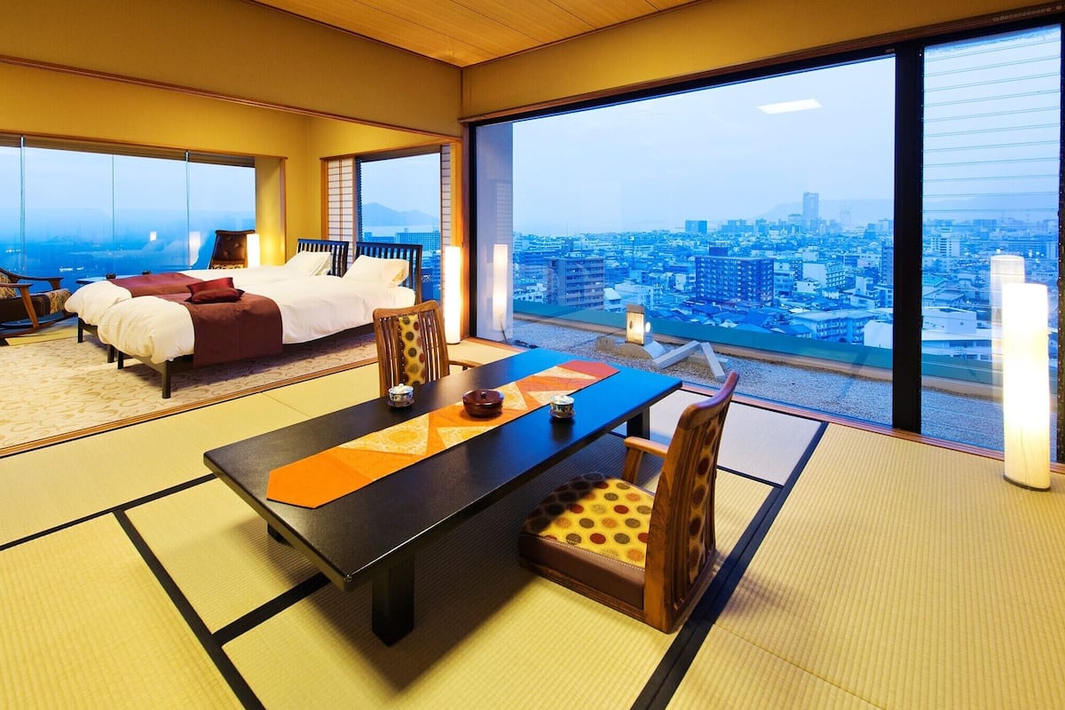 A hotel room with tatami flooring, a dining table, a sofa, and a large window with a city view.