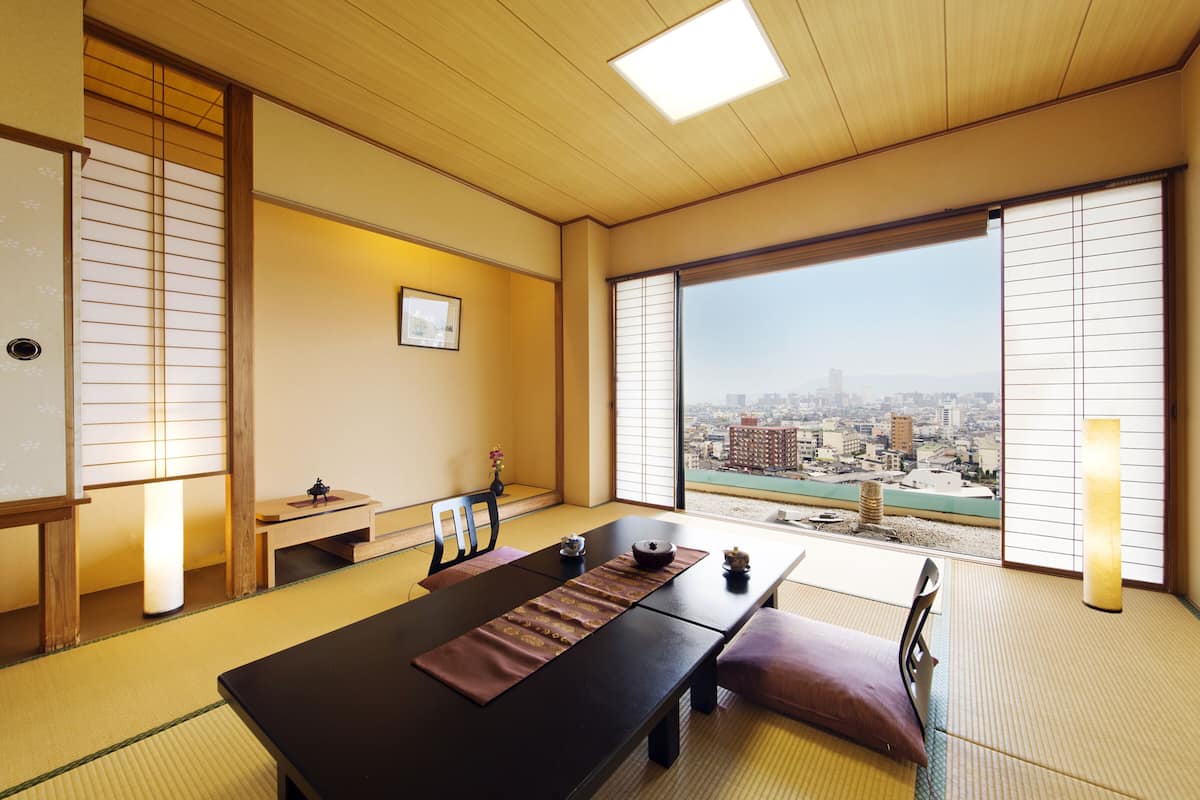 Una habitación japonesa tradicional con suelo de tatami, una mesa baja con un felpudo oscuro, puertas correderas shoji y vistas a la ciudad a través de amplios ventanales.
