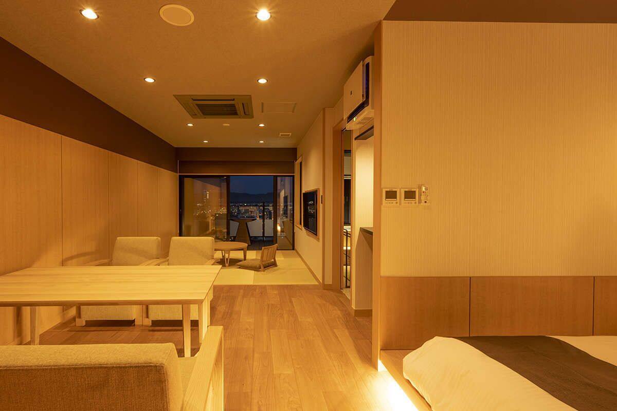 Habitación de hotel con piso de madera, una cama, un escritorio y una silla. Hay una ventana con vistas a la ciudad de noche.