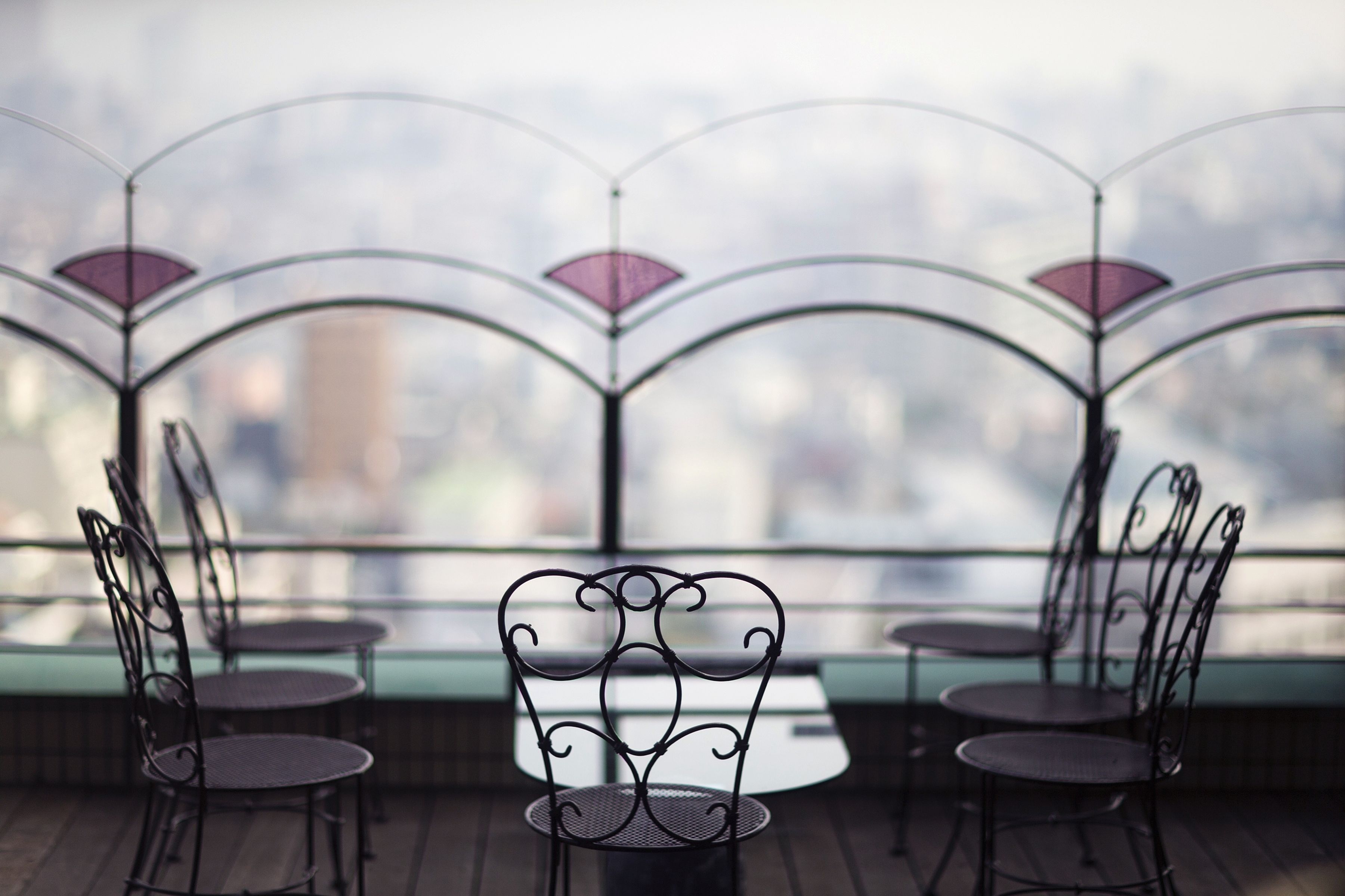 Una terraza con sillas y mesas de metal, barandilla de vidrio y vista panorámica de la ciudad.