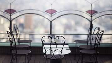Una terraza con sillas y mesas de metal, barandilla de vidrio y vista panorámica de la ciudad.