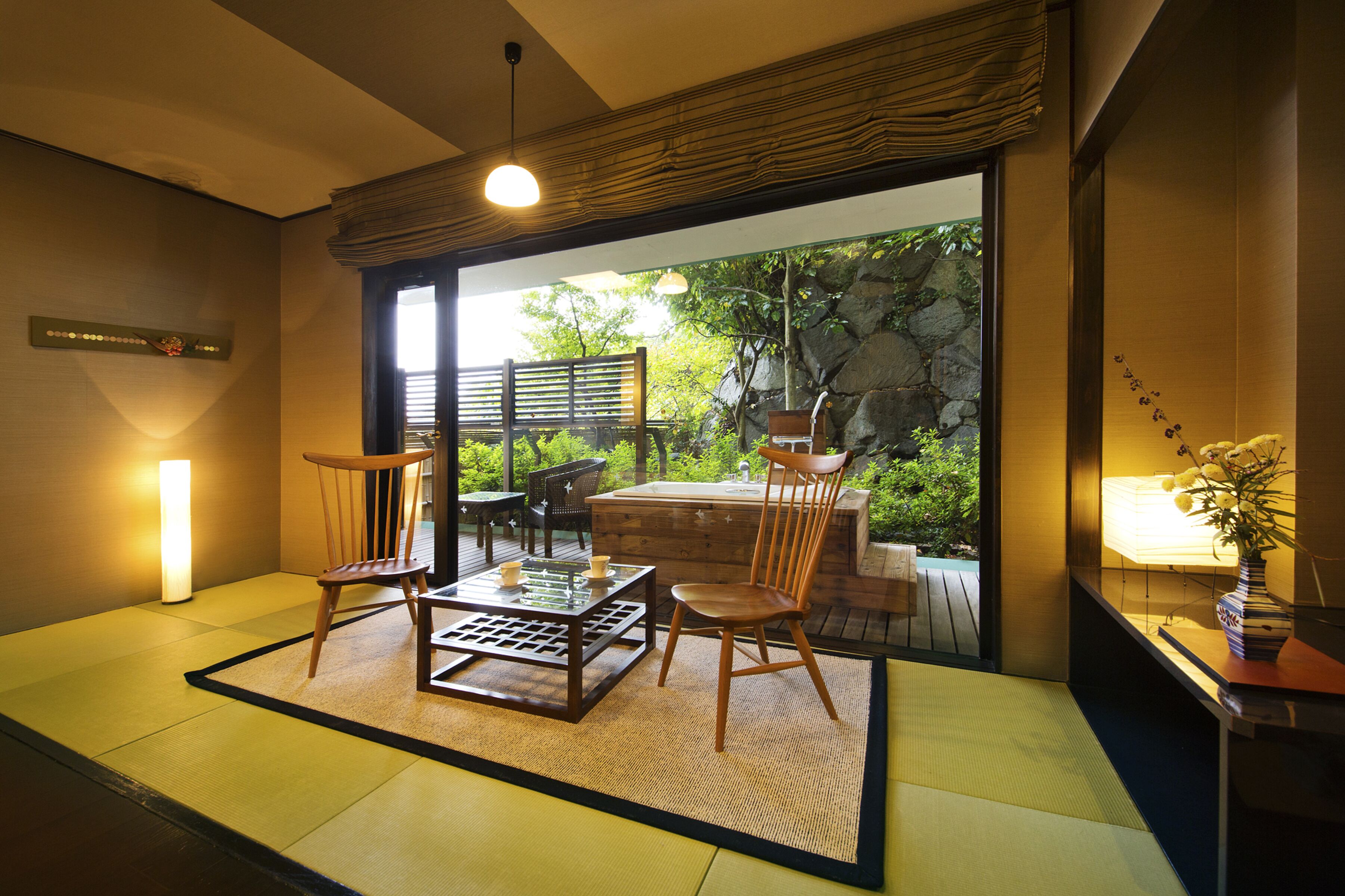 Ein traditioneller japanischer Raum mit Tatami-Böden, einem niedrigen Tisch, Stühlen und Blick auf einen Garten durch eine Schiebetür.