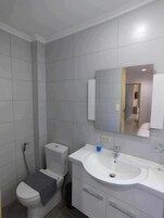 A modern bathroom with grey tiles, a white toilet, a sink with a mirror above it, and a shower area.