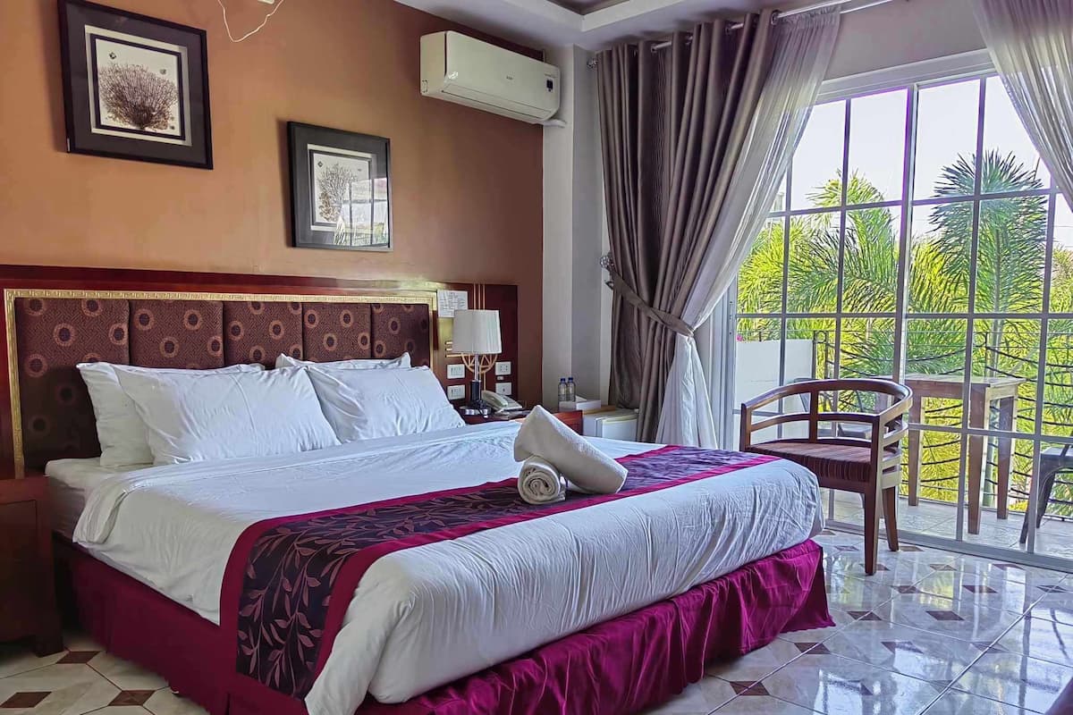 A hotel room with a large bed, a chair, a balcony with a view of greenery, and an air conditioning unit on the wall.
