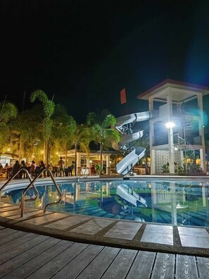 Una piscina con un tobogán moderno, rodeada de palmeras y un edificio con una escalera de caracol.