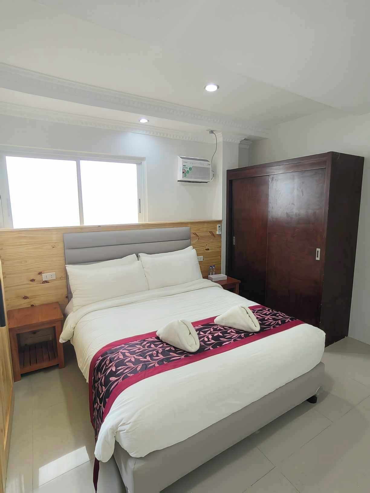 A hotel room with a bed, wooden headboard, bedside tables, a wardrobe, and an air conditioning unit.