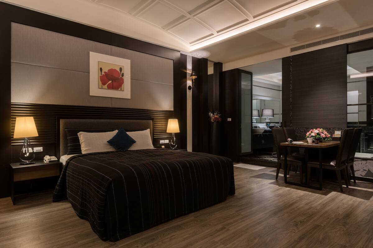 Ein modernes Hotelzimmer mit einem großen Bett, Nachttischlampen, einem Schreibtisch mit Stuhl und Blick auf den Küchenbereich.