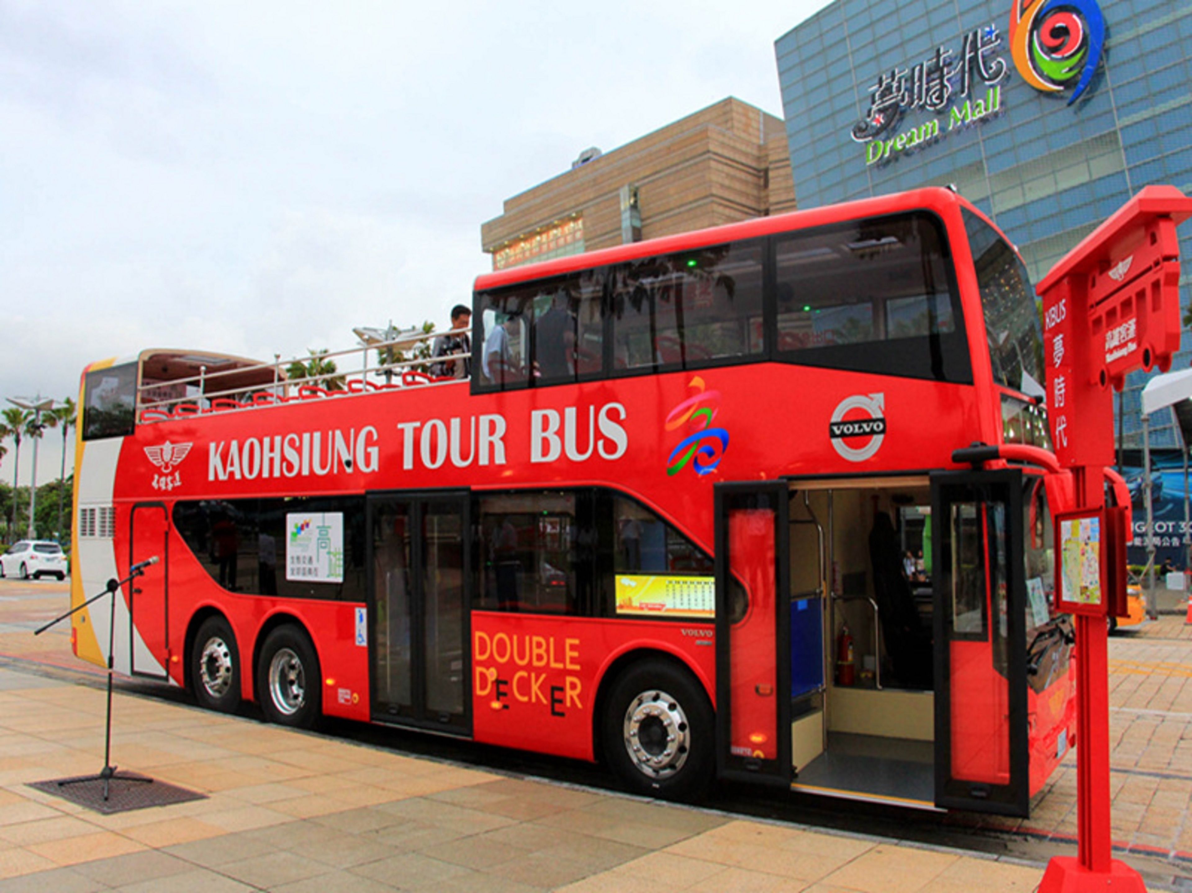 Un autobús turístico rojo de dos pisos con la inscripción "Kaohsiung Tour Bus" y "Doble Piso", estacionado frente a un edificio con el letrero "Dream Mall".