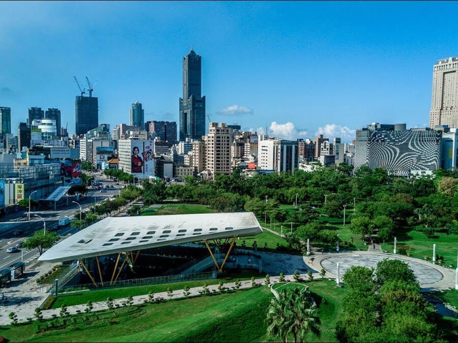 A modern cityscape with a prominent white building featuring a unique roof design, surrounded by lush green parks and a variety of skyscrapers.