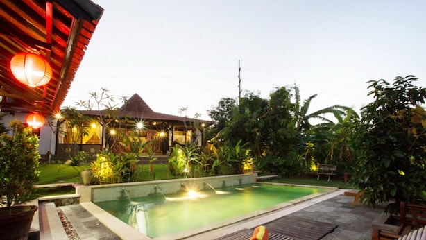 A swimming pool with a wooden deck and lounge chairs, surrounded by lush greenery and a traditional building with a thatched roof. - Kubu Ampo Villa (Kerobokan)