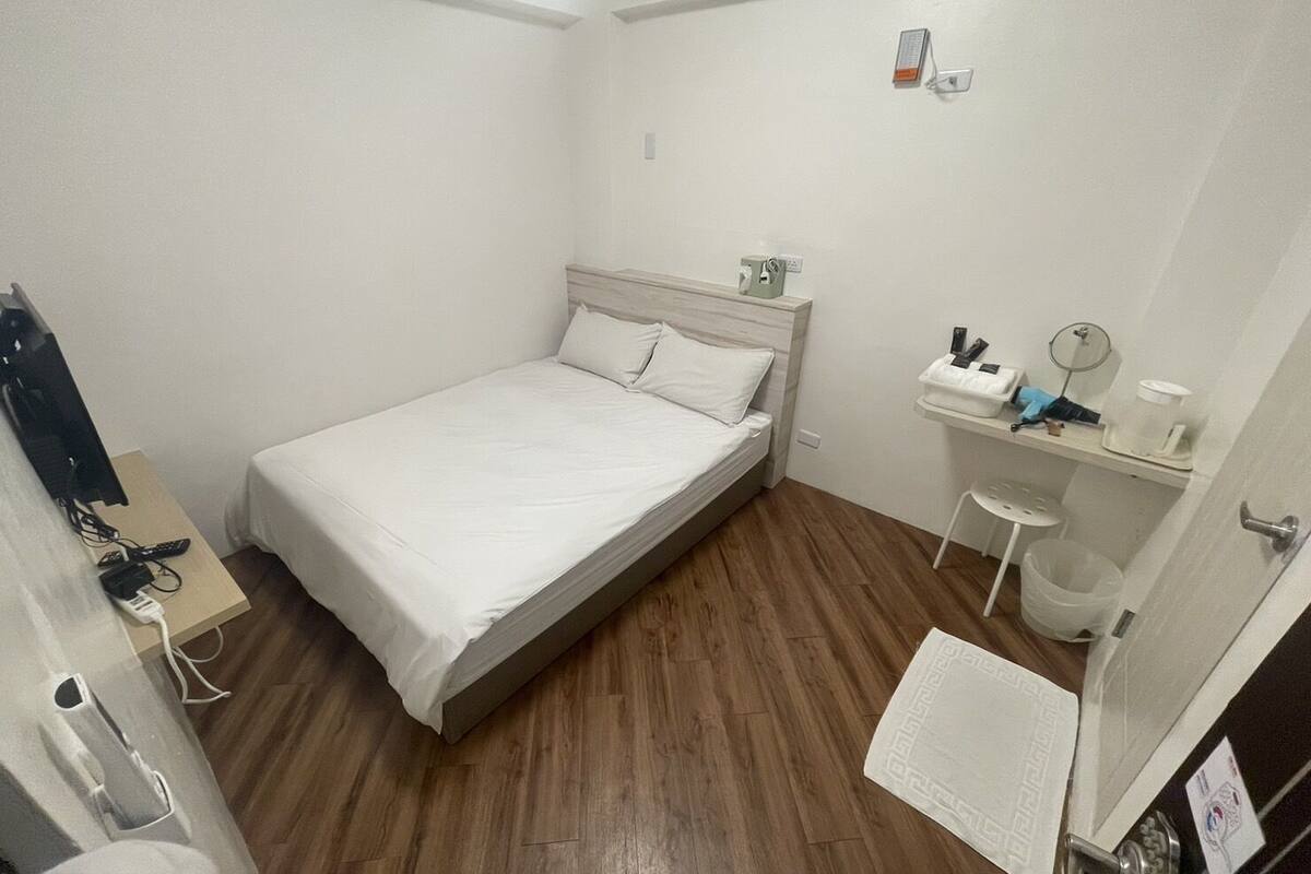 A small, neatly arranged bedroom with a single bed, a bedside table, a small shelf with toiletries, and a wooden floor.