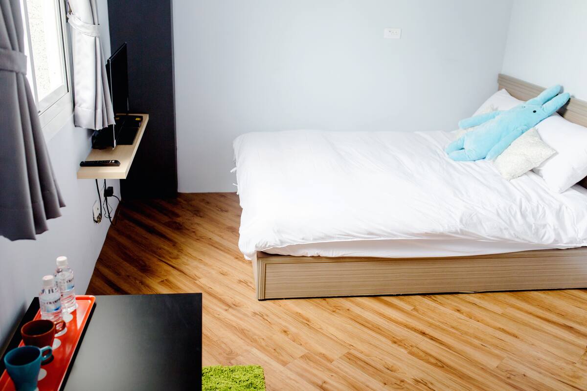 A neatly made bed with white bedding and a wooden headboard, a small bedside table with a remote control and a water bottle, and a window with curtains.