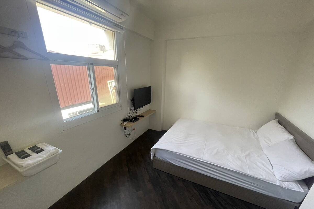 A small, clean room with a bed, a TV on a shelf, a window, and a small table with a phone.