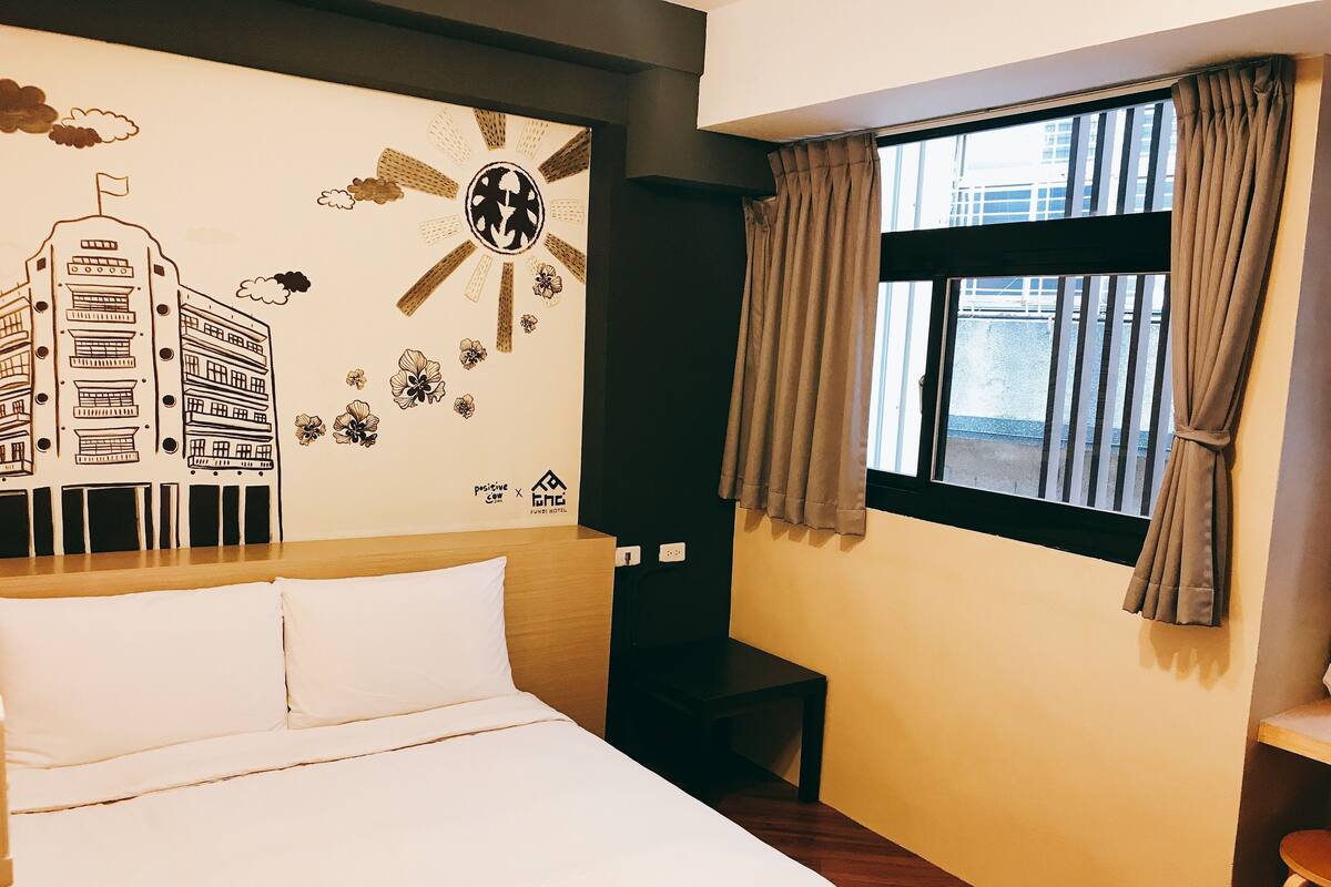 A hotel room with a bed, a wall mural, a window with curtains, and a small table.