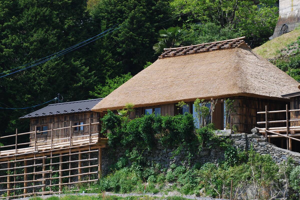 Une maison traditionnelle en bois, avec un toit de chaume, entourée d’une végétation luxuriante et d’un mur de pierre.