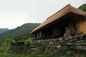 A rustic wooden cabin with a thatched roof, situated on a stone wall with a view of lush green hills in the background.