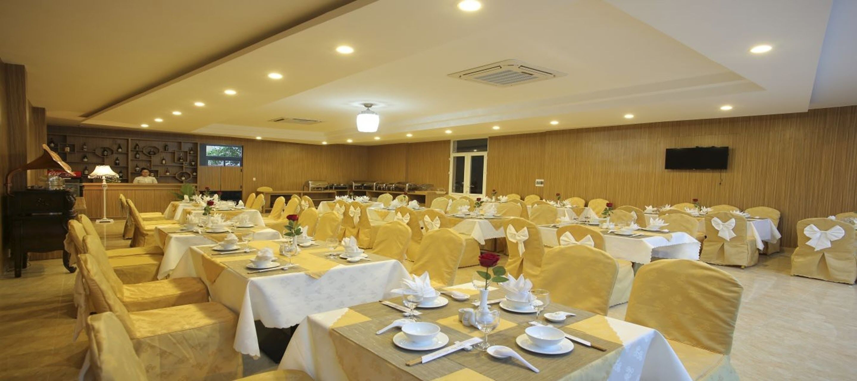Une salle de banquet avec des tables dressées pour un événement formel, agrémentée d’un mur recouvert de panneaux de bois et d’un téléviseur à écran plat fixé au mur.
