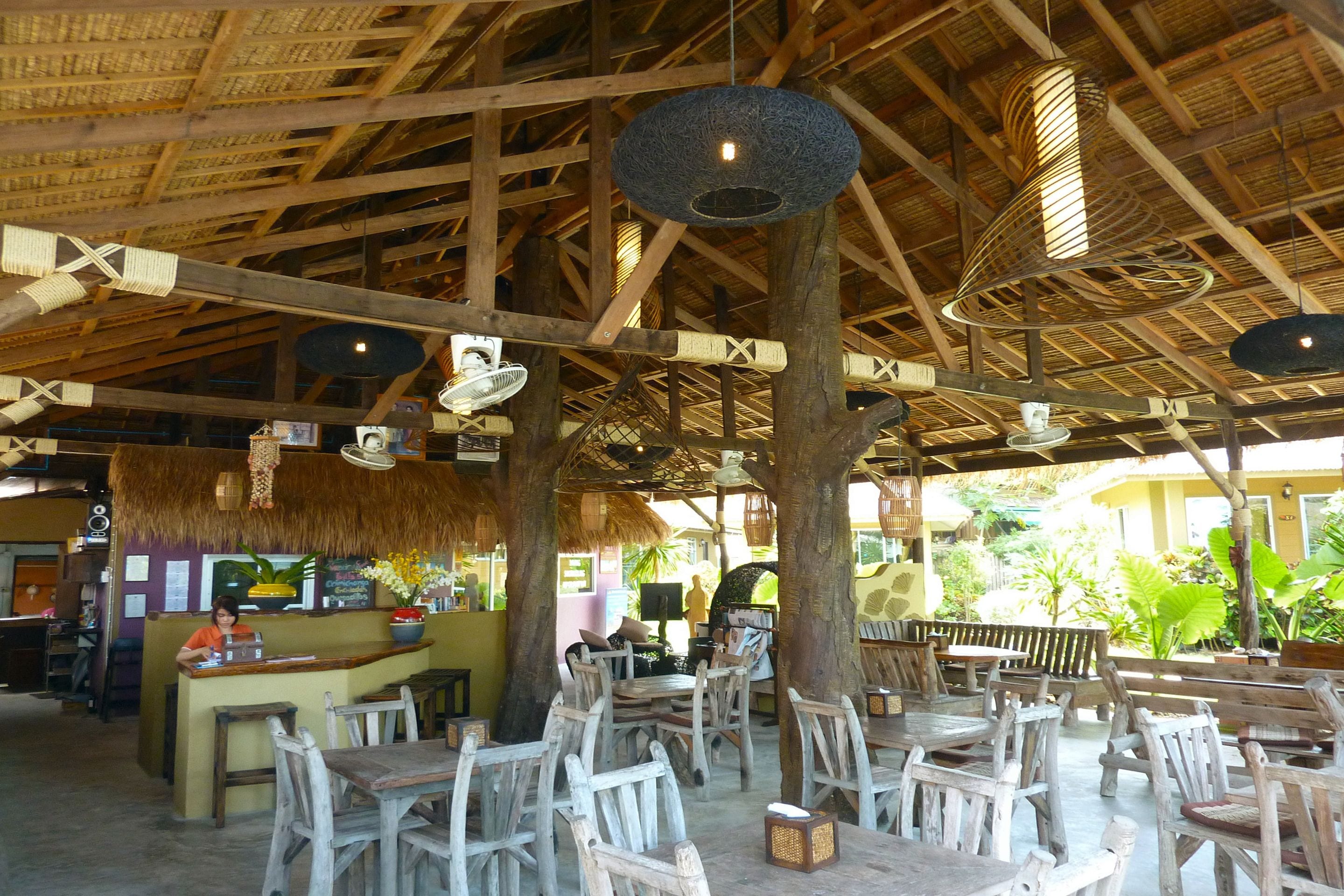 An indoor dining area with wooden tables and chairs, a thatched ceiling, and hanging light fixtures.