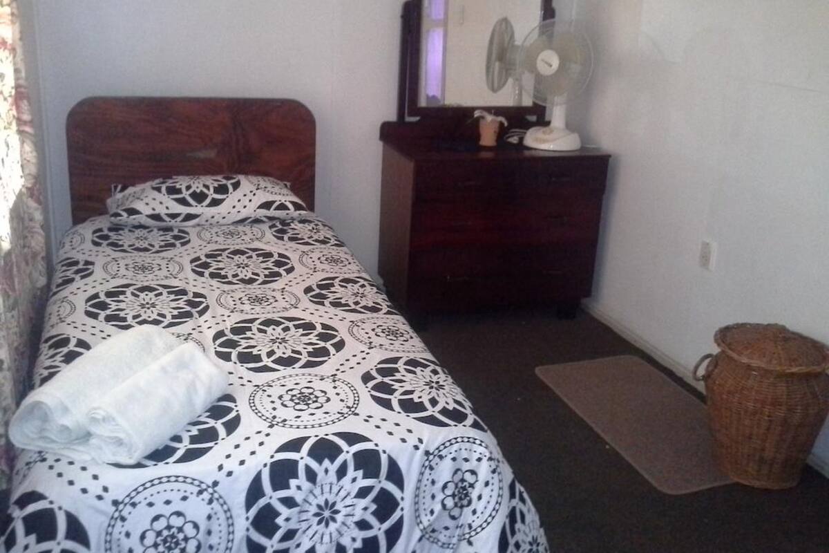 A bedroom with a bed, a dresser, a mirror, a fan, and a waste basket.