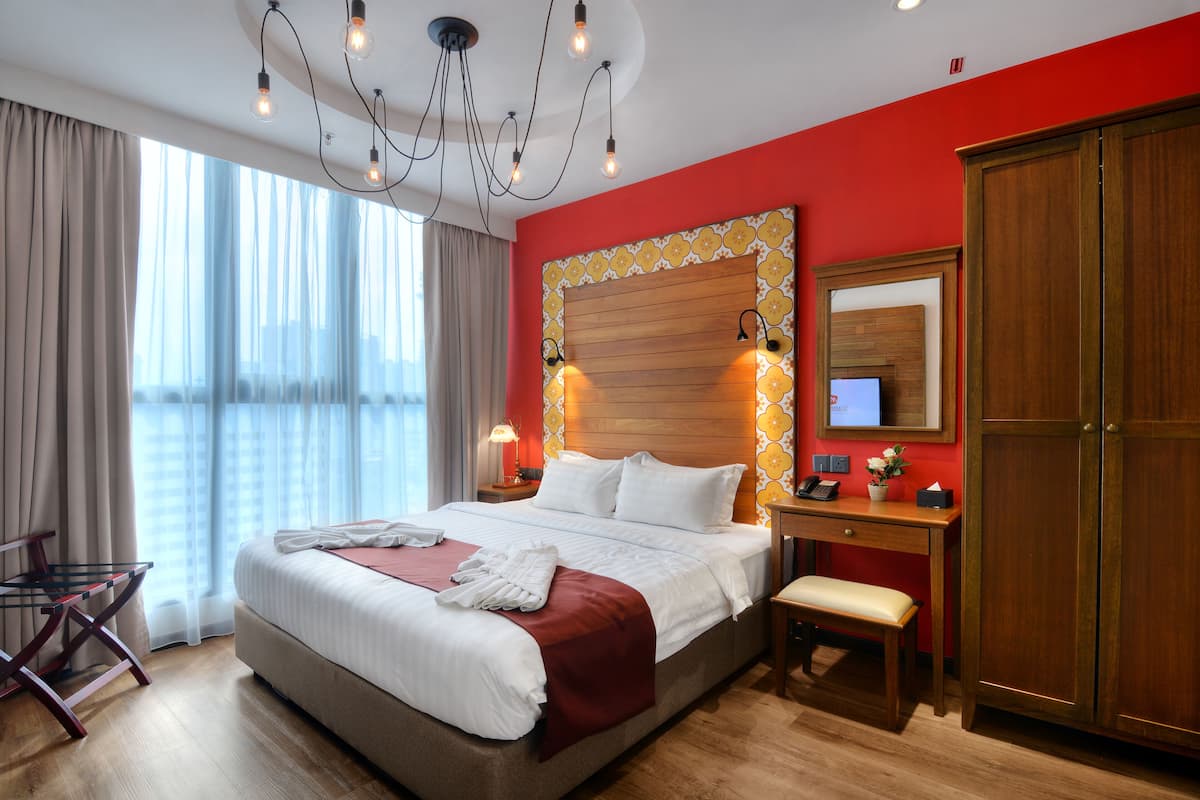 A hotel room with a large bed, a desk with a chair, a wardrobe, and a large window with curtains.
