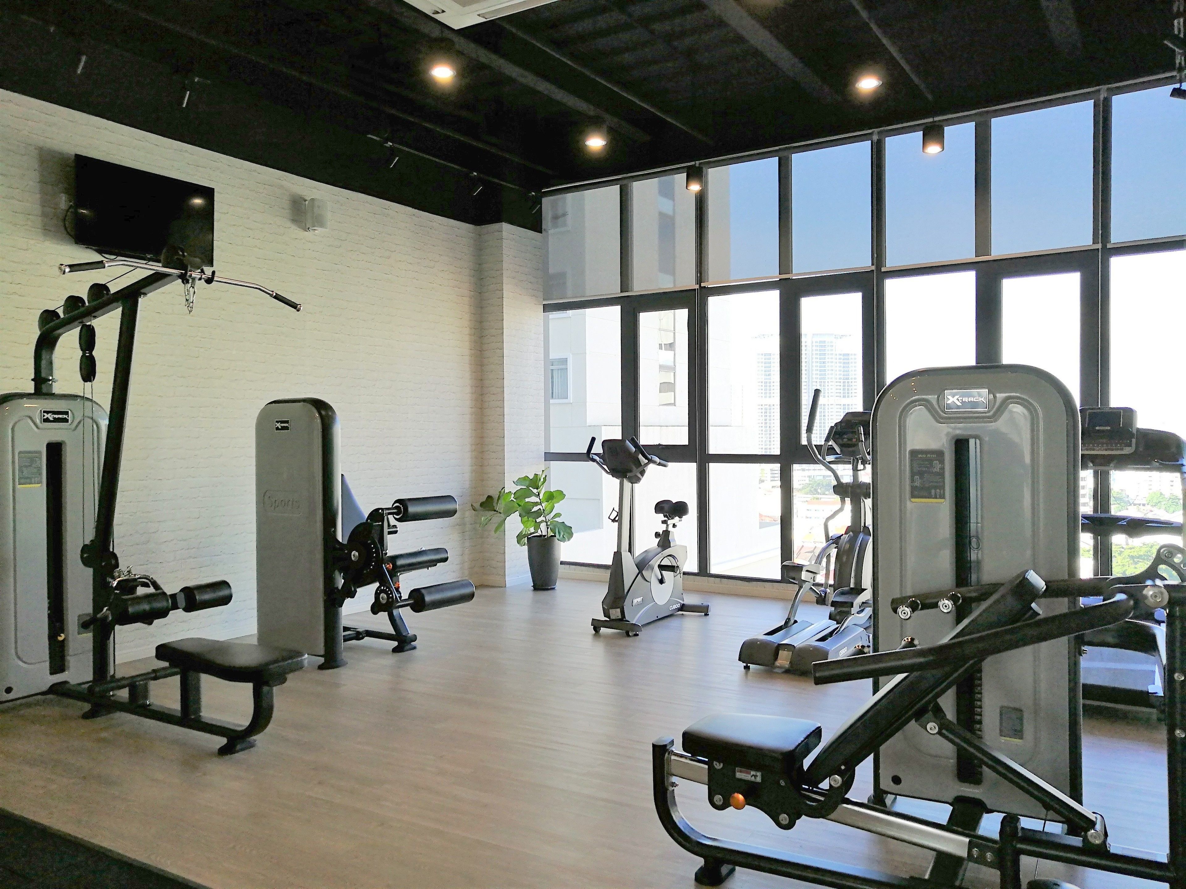 A gym with various exercise machines, a television on the wall, and large windows offering a view of the city.